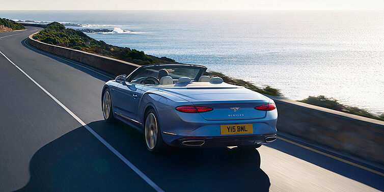 Bentley Abu Dhabi New Bentley Continental GTC Azure convertible rear view in Light Windsor blue paint with 22 inch ten spoke wheels driving dynamically on an ocean road in the sunshine